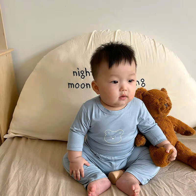 Baby in Snuggle Bear Pajamas sitting on bed with teddy bear.