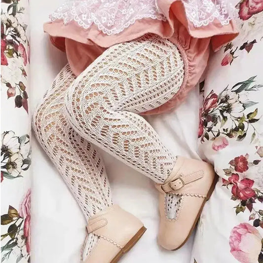 Baby wearing hollow out mesh tights with a lace dress and pink shoes, lying on a floral-patterned blanket.